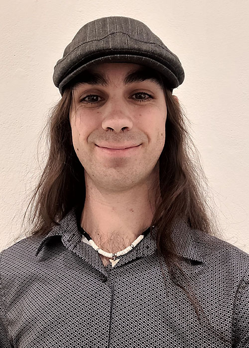 Headshot of a young man with long dark hair wearing a grey shirt and a striped cap.